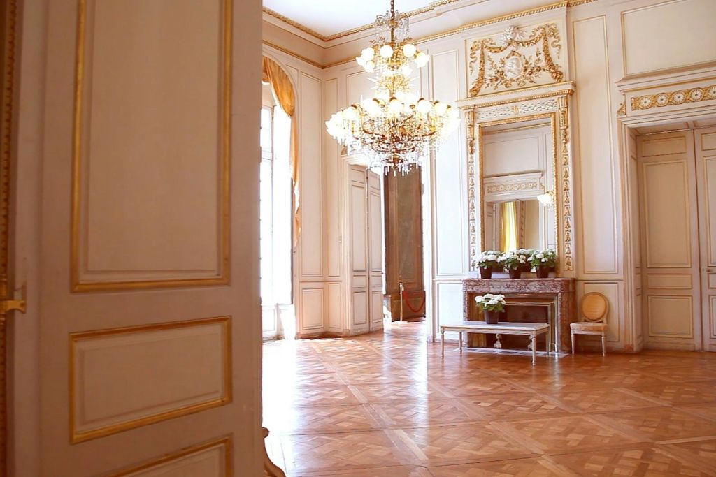 샹들리에가 있는 화려한 객실, 벽난로 위의 커다란 거울, 망토에 핀 꽃, 쪽모이 세공 마룻바닥. 커튼이 있는 햇빛이 들어오는 창문.