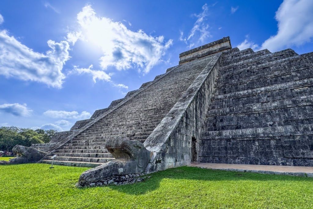 구름이 낀 푸른 하늘을 배경으로 뱀의 조각품이 있는 계단이 있는 석조 피라미드. 녹색 잔디가 바닥을 둘러싸고 있습니다.