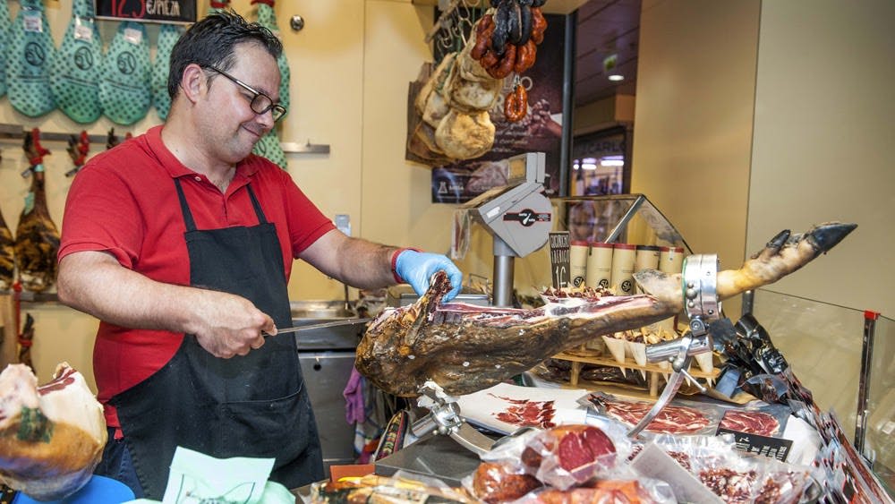 빨간색 셔츠와 검은색 앞치마를 입은 사람이 델리에서 고기를 잘게 썰고, 고기와 제품을 배경으로 매달려 있습니다.