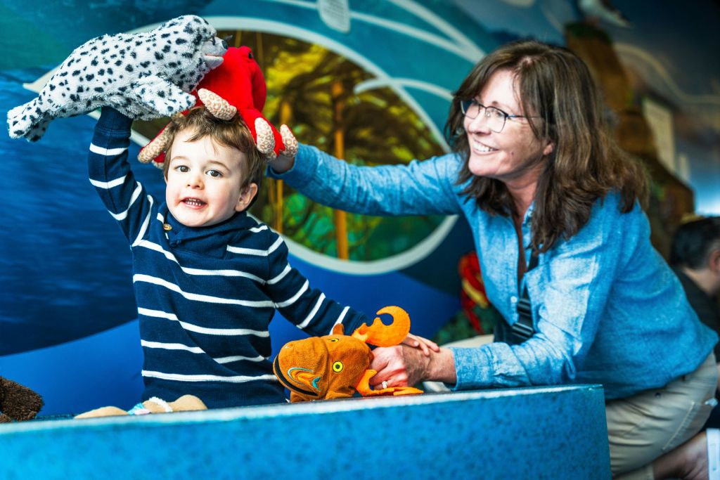 줄무늬 셔츠를 입은 어린이가 어른이 미소를 지으며 박제 동물을 안고 화려한 분위기에서 머리에 푹신한 장난감을 하나 더 맞춥니다.