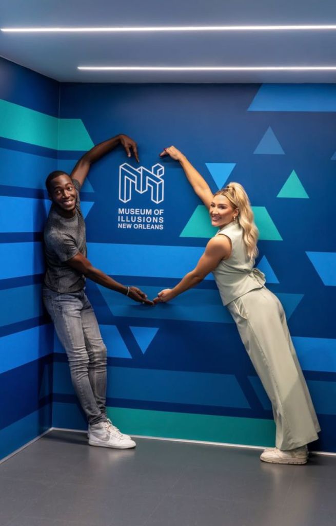 "Museum of Illusions New Orleans" 라는 글과 함께 벽 근처에서 장난스럽게 포즈를 취하는 두 사람. 하나는 로고를 가리키고 있습니다.