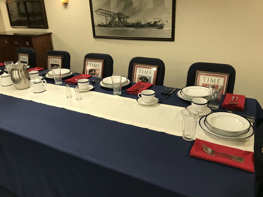 흰색과 파란색 침구, 접시, 컵, 식기류, 빨간색 냅킨이 놓인 테이블 세트. 의자에는 Time 잡지 표지가 액자에 담겨 있습니다.