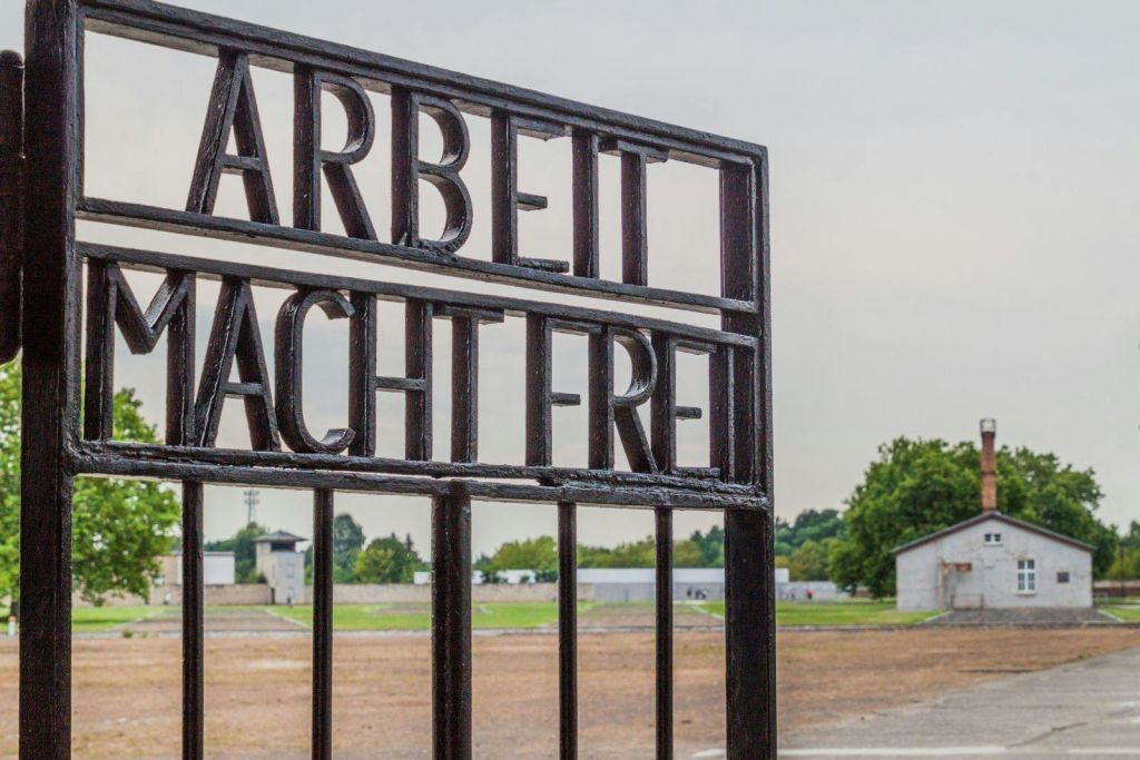 "Arbeit Macht Frei" 문구가 새겨진 연철문으로, 건물과 나무가 멀리 보이는 황량하고 탁 트인 지역이 내려다보입니다.