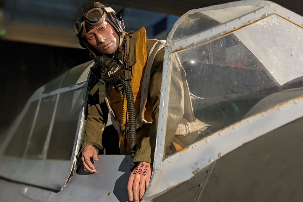 제2차 세계대전 조종사로 포즈를 취하고 있는 마네킹이 조종석에 앉아 고글과 헤드셋을 착용하고 있습니다. 장갑에는 손으로 쓴 메모가 새겨져 있습니다.