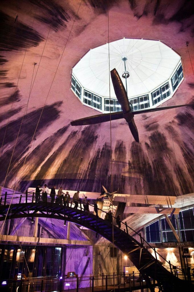 중앙 조명 기구와 눈에 띄는 구조 대들보가 있는 커다란 돔 천장 아래 높은 다리를 걷고 있는 사람들.