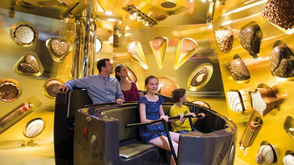 벽면을 배경으로 초콜릿 장식이 돋보이는 놀이공원 어트랙션을 4인 가족이 타고 있습니다.
