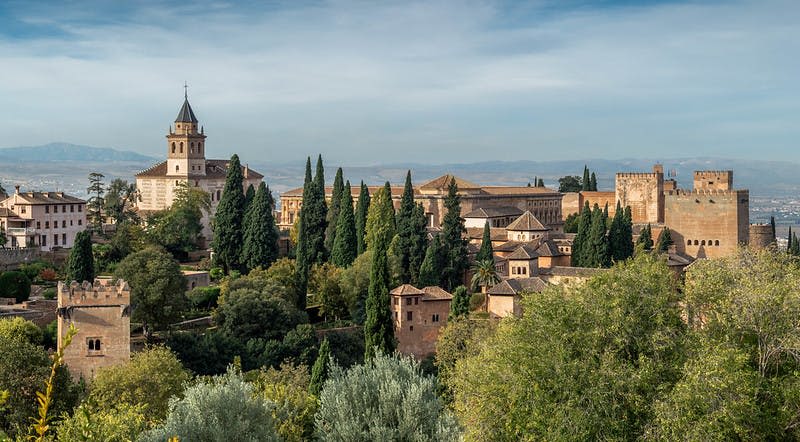 높은 편백나무, 소박한 건물, 부분적으로 흐린 하늘 아래 먼 산맥이 있는 역사적인 단지의 경치.