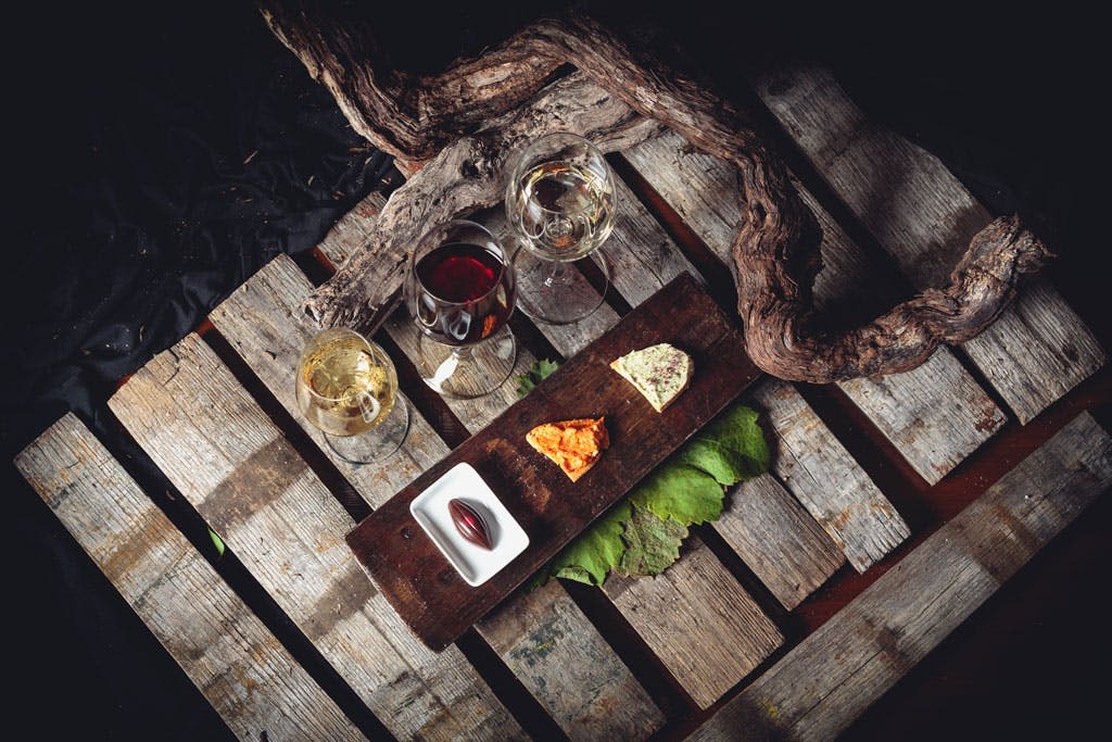 덩굴이 우거진 소박한 나무 표면에 세워진 나무판에 레드, 치즈, 초콜릿 중 하나인 화이트 와인 두 잔.