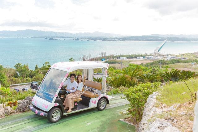 푸른 물과 녹음이 우거진 해안 풍경이 내려다보이는 하트로 장식된 골프 카트를 타고 있는 두 사람.