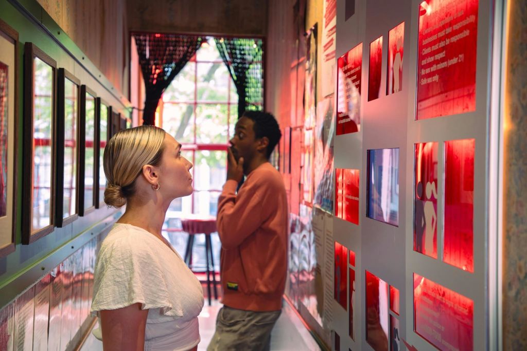 두 사람은 좁고 조명이 밝은 갤러리 공간에서 액자에 담긴 예술품과 벽 텍스트를 관찰하고 있으며, 끝에는 커다란 창문이 있습니다.
