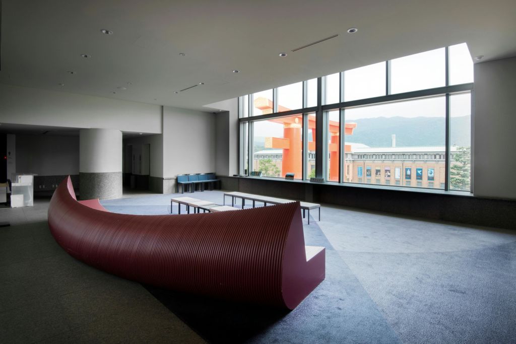 커다란 창문이 있는 넓은 객실로 곡선형 마룬 벤치, 테이블, 의자, 외부가 보이는 주황색 건물이 있습니다.