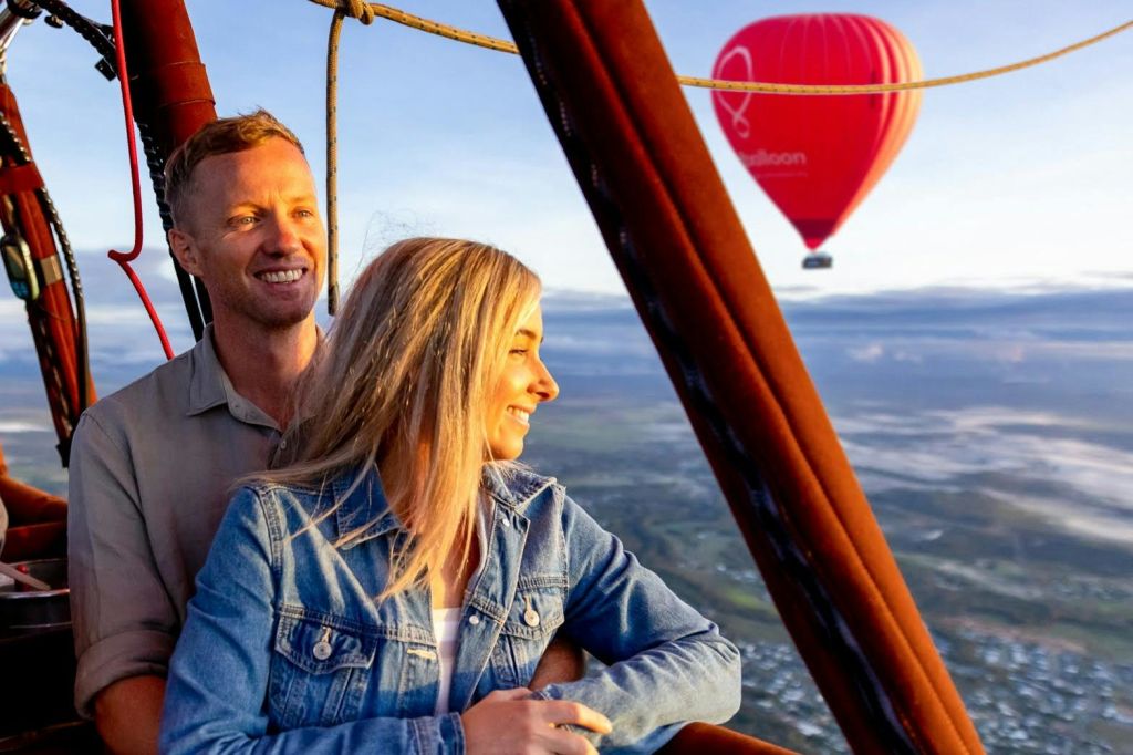 아래에 경치가 아름다운 열기구 바구니에 서 있는 한 남녀가 미소를 짓고 있습니다.