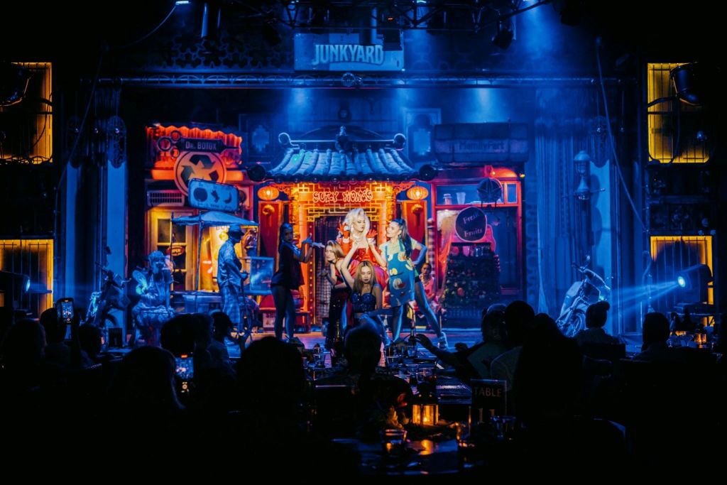 무대에서 극적인 조명으로 빛나는 공연자와 테이블에 앉아 있는 관객이 있는 활기찬 나이트클럽 장면.