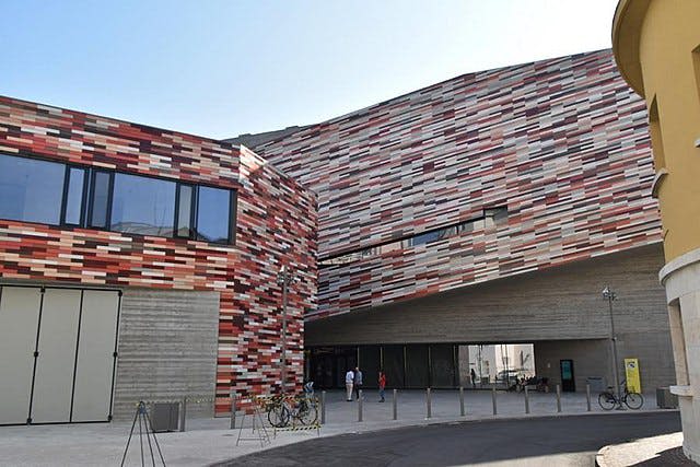 화려한 패턴의 외관, 커다란 창문, 개방형 출입구를 갖춘 현대적인 건물입니다. 세 사람과 여러 대의 자전거가 보입니다.