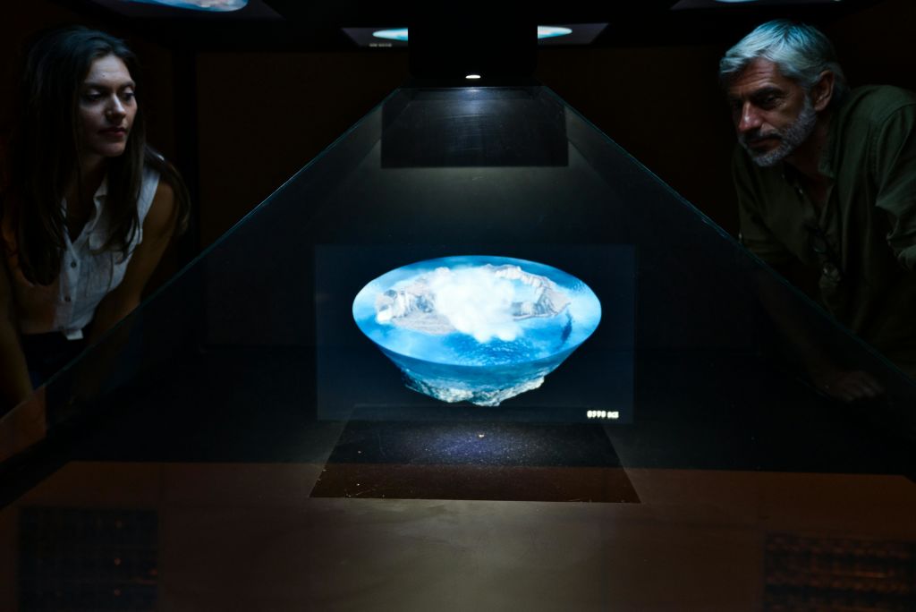 한 여성이 어두운 방 안에서 물과 구름이 떠다니는 섬의 홀로그램 디스플레이를 관찰하고 있습니다.