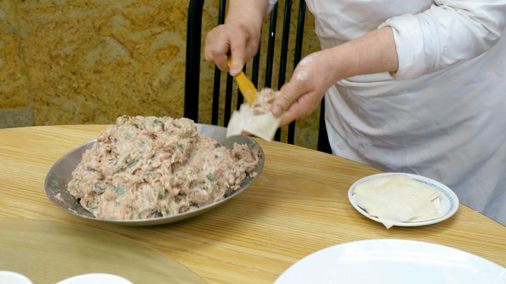 한 사람이 주걱으로 만두를 만들어 반죽 포장지에 채운 고기를 나무 테이블 위에 올려놓습니다.