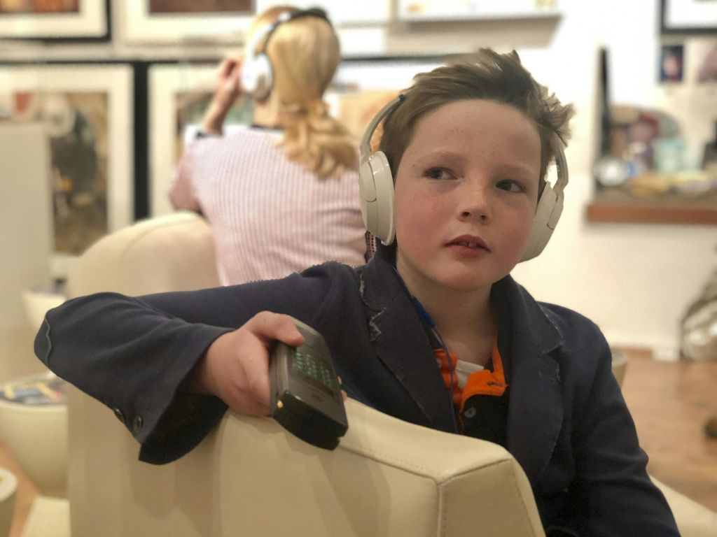 헤드폰을 든 어린 소년이 실내에 앉아 리모컨을 들고 있습니다. 배경의 성인도 헤드폰을 착용합니다.