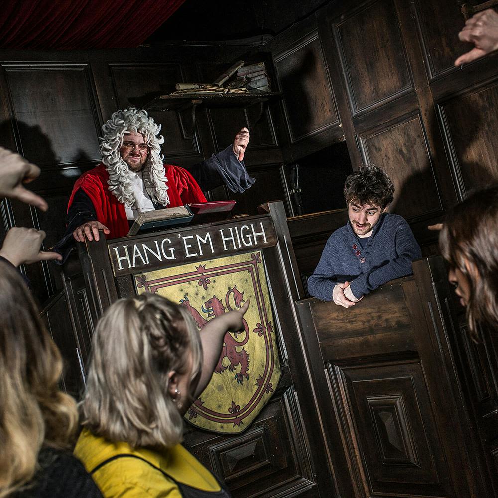 흰 가발과 빨간 가운을 입은 판사가 사람들이 가리키고 있는 증인 스탠드에서 한 남자를 주재합니다. 표지판에는 "Hang 'em High" 라고 쓰여 있습니다.