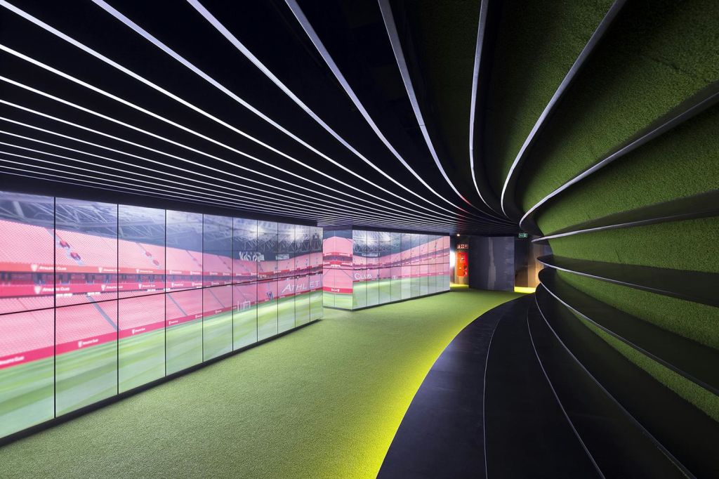 곡선형 벽, 빨간색 경기장 이미지가 표시된 디지털 스크린, 머리 위로 줄무늬 조명이 있는 녹색 바닥이 있는 현대적인 복도입니다.