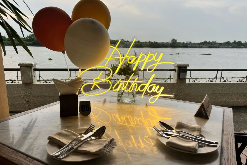 강 옆에 있는 풍선, 식기류, 냅킨, 테이블 번호 06이 있는 식탁에 "생일 축하합니다" 네온 사인이 있습니다.