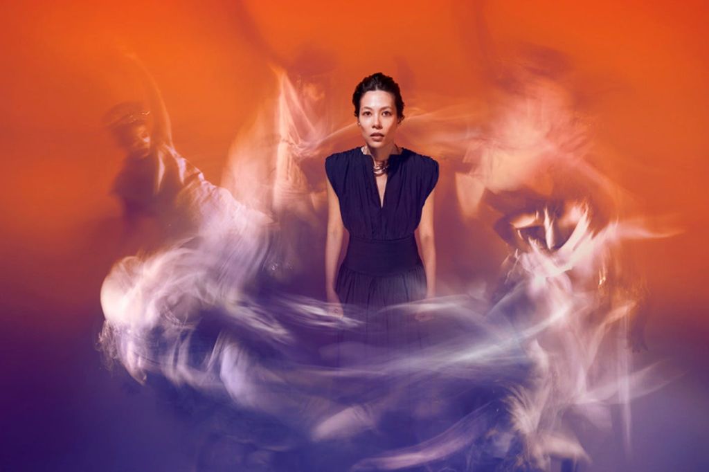 어두운 드레스를 입은 한 여성이 추상적인 밝은 산책로와 주변의 흐릿한 형상이 있는 활기찬 주황색 배경에 서 있습니다.