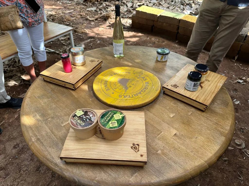 나무 쟁반에 와인, 항아리, 음식이 놓인 둥근 나무 테이블. 테이블 근처에 서 있는 두 사람이 부분적으로 보입니다.