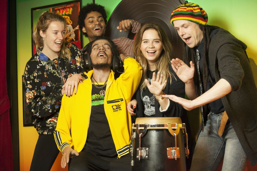 다섯 명이 실내에서 함께 음악을 즐기고, 한 명은 드럼을 연주하고, 다른 한 명은 헤드폰을 씁니다. 그들은 활기차고 즐겁게 보입니다.