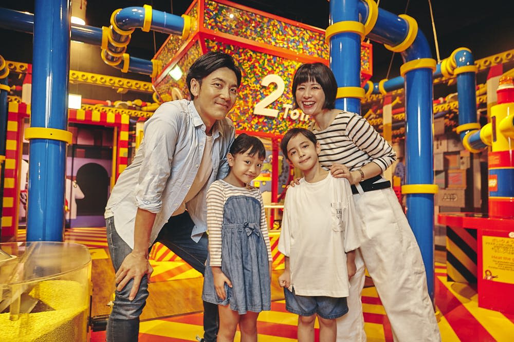 튜브, 액티비티, 커다란 '2' 가 배경으로 보이는 다채로운 실내 놀이 공간에서 미소를 짓고 있는 4인 가족.