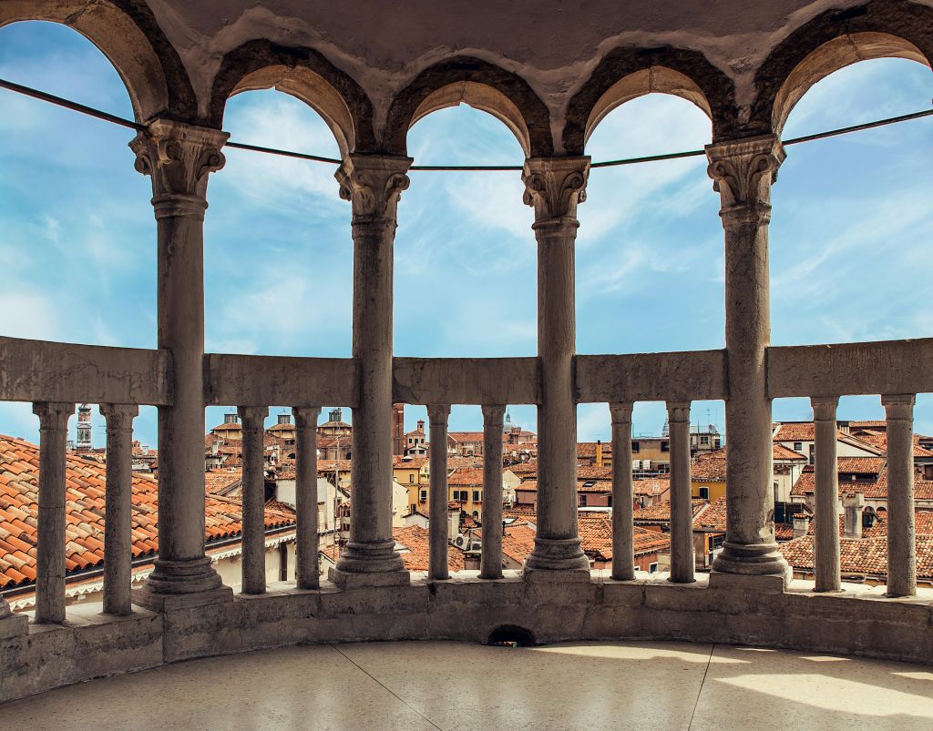 부분적으로 흐린 푸른 하늘 아래 붉은 지붕의 건물이 있는 도시 풍경이 내려다보이는 아치형 기둥이 있는 발코니.