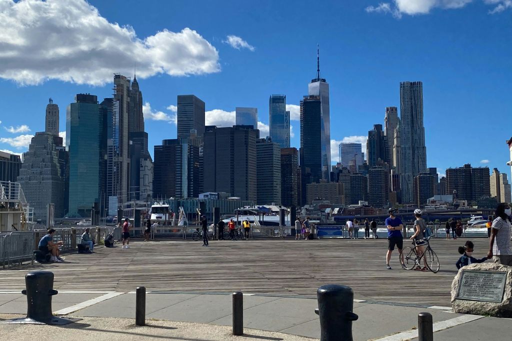 푸른 하늘 아래 높은 건물의 스카이라인, 구름, 산책로에서 걷고 자전거 타는 사람들, 물가에 정박한 보트.