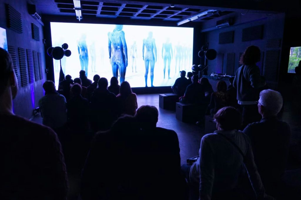 파란색으로 추상적인 휴머노이드 형상을 보여주는 대형 스크린을 마주하고 있는 조명이 희미한 방에 앉아 있는 사람들.