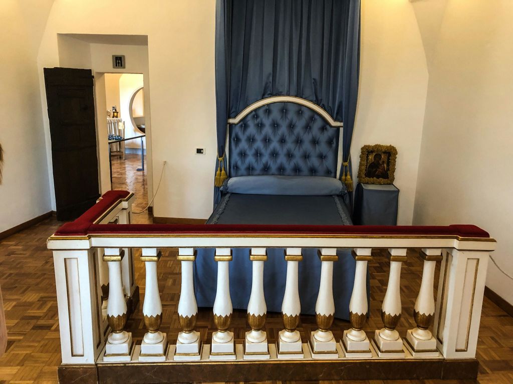 파란색 캐노피 침대, 화려한 흰색과 금색 난간, 나무 바닥, 침대 옆 테이블에 작은 액자 그림이 있는 고급스러운 객실입니다.