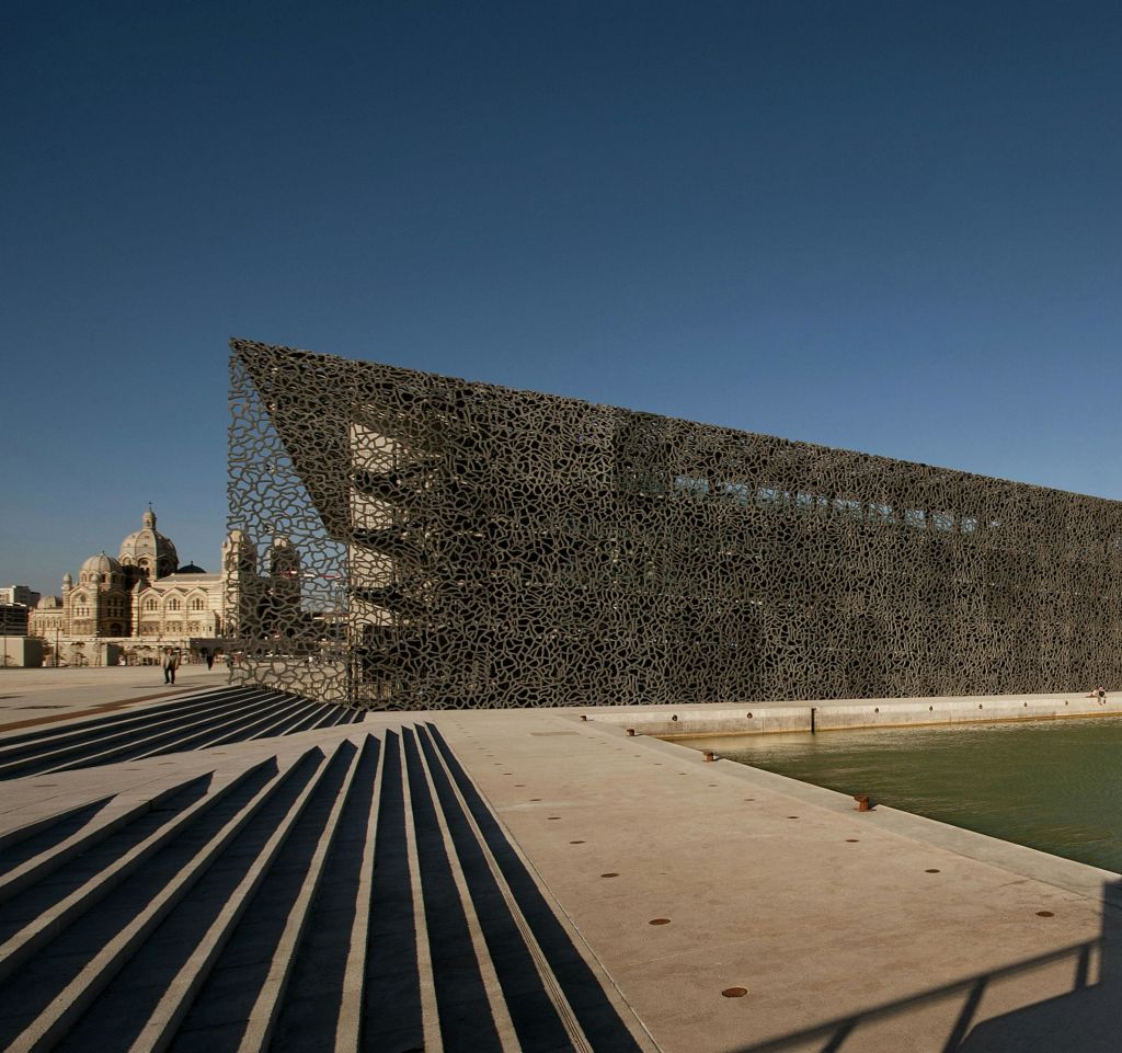 수체 옆의 정교한 격자 디자인이 돋보이는 현대적인 건물. 역사적인 건물을 배경으로 계단과 소수의 사람들이 앞에 서 있습니다.