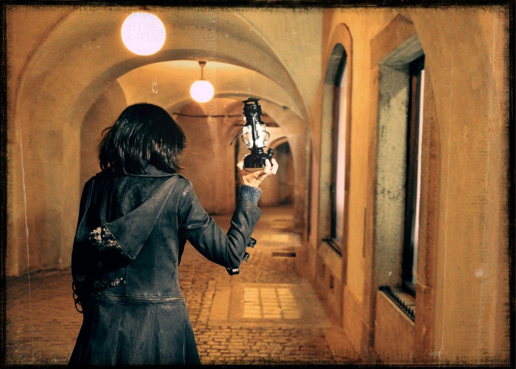 어두운 재킷을 입은 사람이 돌담과 걸이식 조명이 있는 아치형의 어두운 복도를 걷고 있습니다.