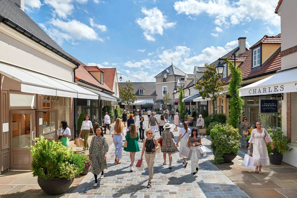 사람들은 부티크, 녹지, 머리 위로 구름이 덮인 푸른 하늘이 있는 고풍스러운 야외 쇼핑 거리를 따라 걷고 쇼핑합니다.