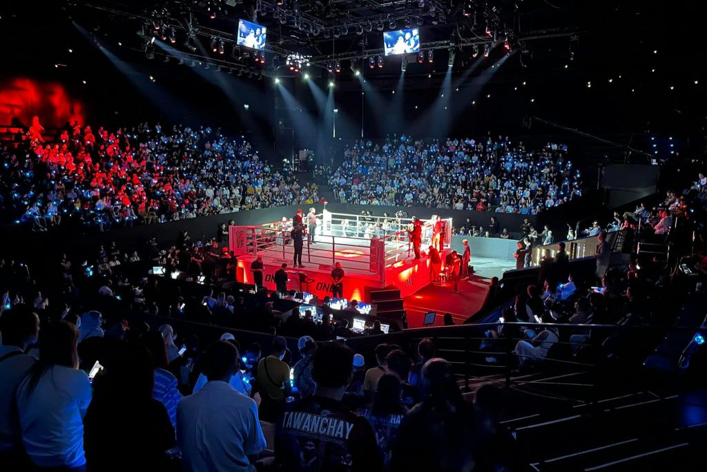 빨간색으로 표시된 복싱 링은 오버헤드 스포트라이트와 대형 스크린이 있는 경기장에서 많은 관객들로 둘러싸여 있습니다.