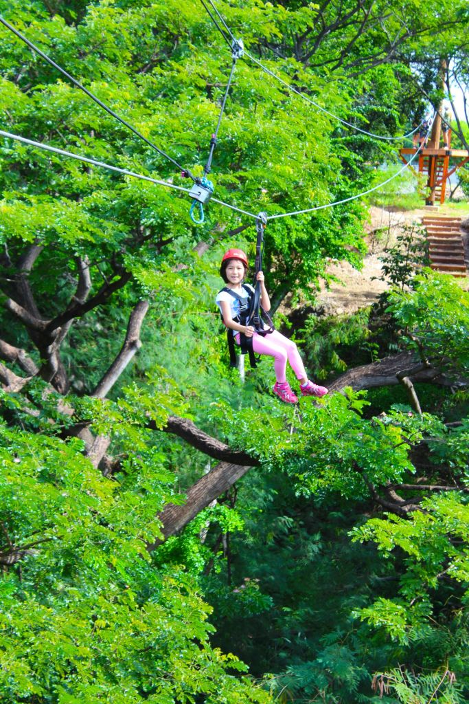 빨간 헬멧과 분홍색 바지를 입고 울창한 녹색 나무로 둘러싸인 짚라인에서 웃고 있는 아이.
