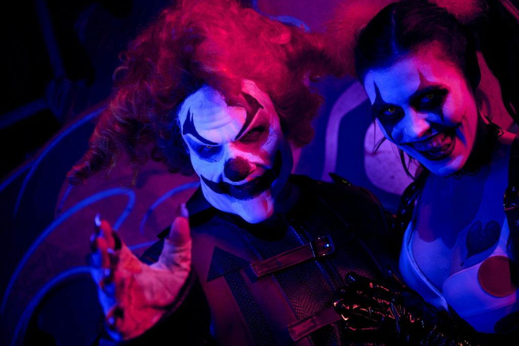 보라색과 파란색 조명으로 빛나는 소름끼치는 광대 메이크업과 의상을 입은 두 사람이 위협적으로 웃고 있습니다.