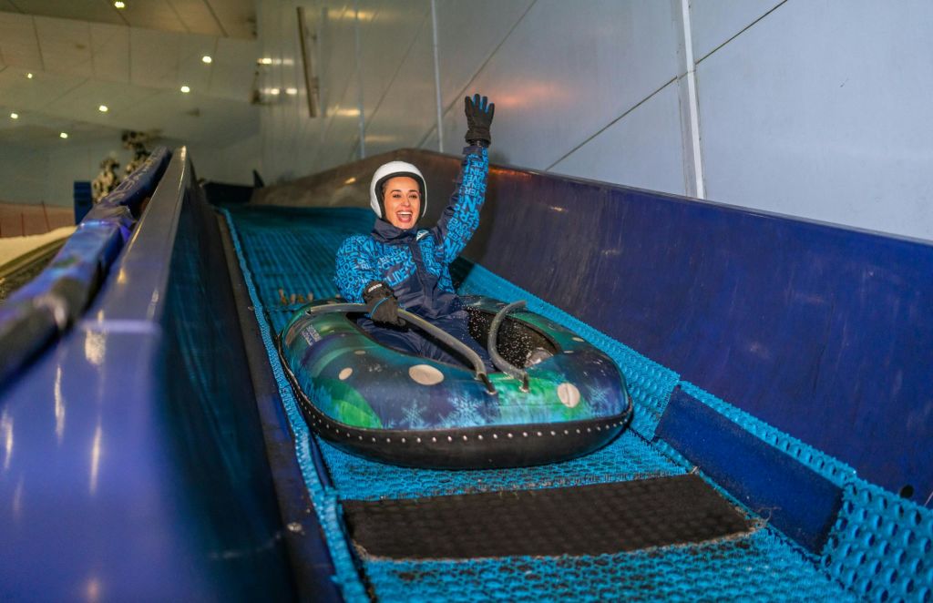 겨울 장비와 헬멧을 쓴 사람이 실내 스노우 튜브 슬라이드를 미끄러지며 웃으며 한 손을 들어 올립니다.