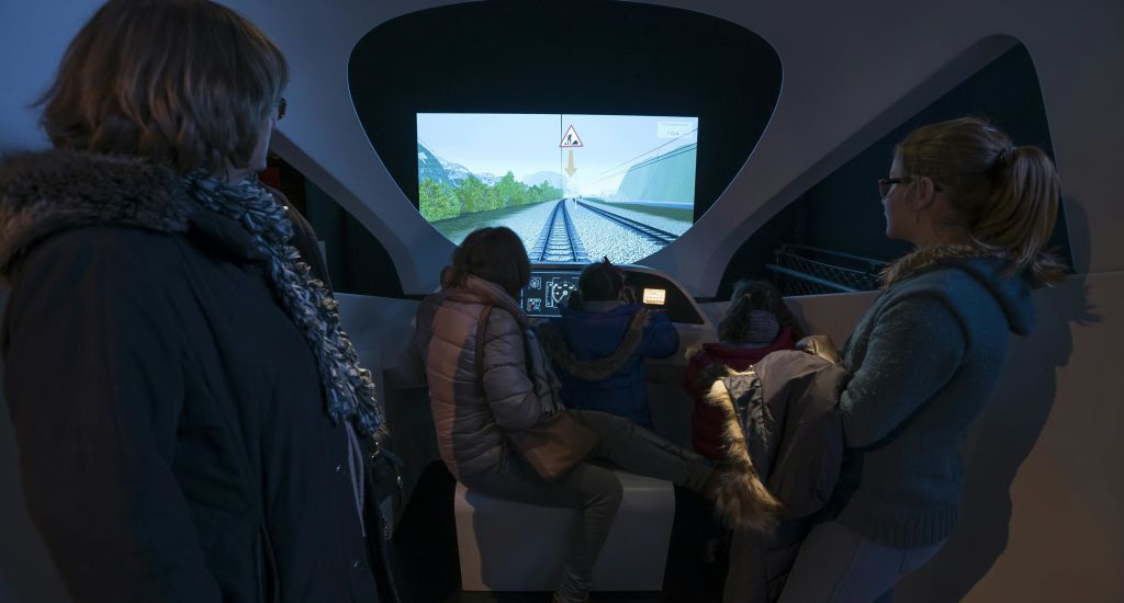 열차 선로와 경고 표지판이 표시된 철도 시뮬레이터 화면과 상호 작용하는 어두운 전시물에 세 사람이 앉아 있습니다.