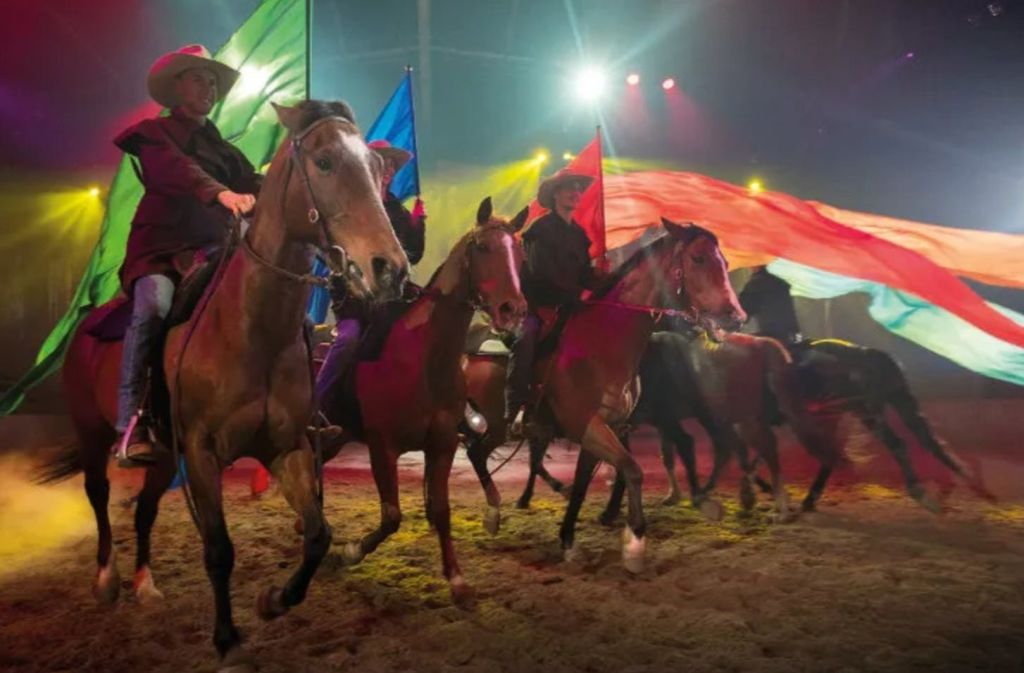 조명이 켜진 경기장에서 화려한 깃발을 들고 승마를 즐기는 승객들.