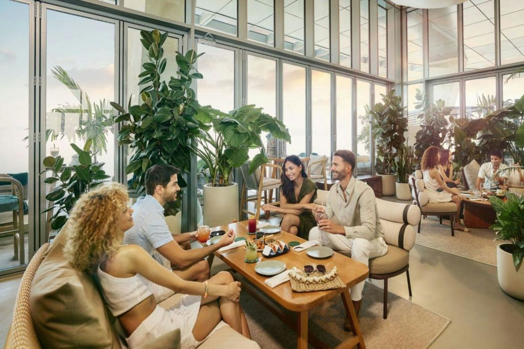 커다란 화분과 유리 벽으로 둘러싸인 현대적인 카페에서 4명이 테이블에 둘러앉아 음료와 간식을 즐기고 있습니다.