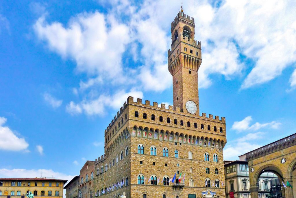 부분적으로 흐린 하늘 아래 높은 시계탑이 있는 중세 스타일의 건물. 아치형 창문의 가운데 줄에 깃발이 걸려 있습니다.