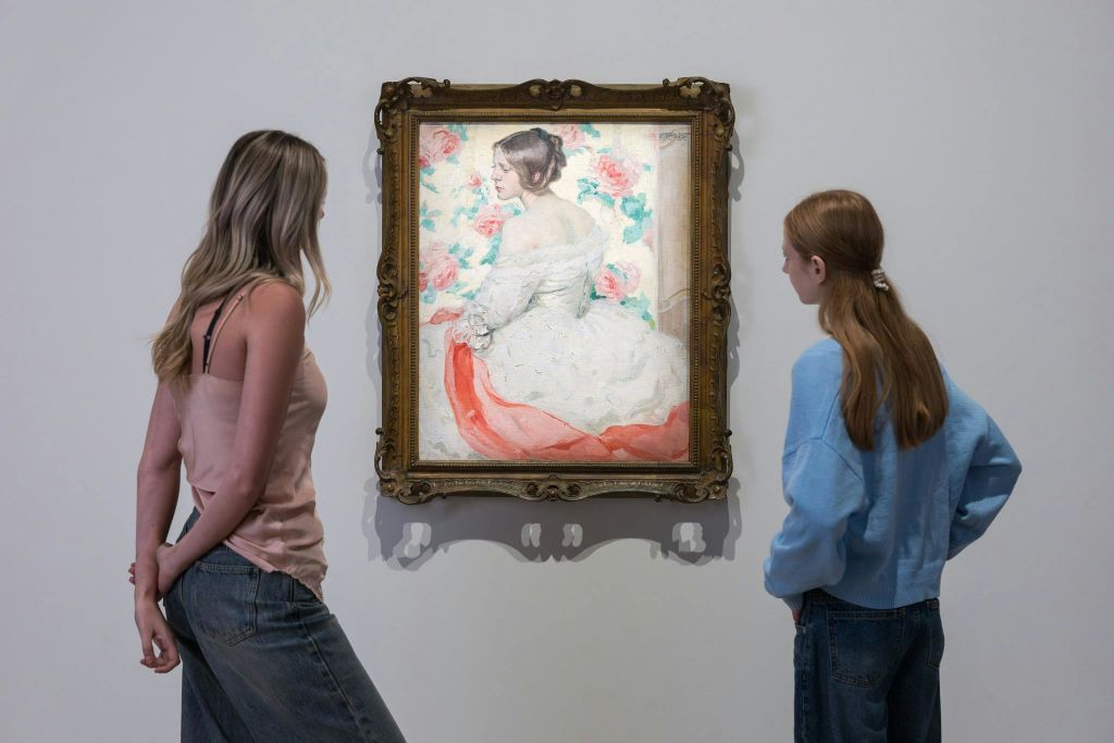 갤러리 벽에 꽃무늬로 둘러싸인 빨간색 띠가 달린 흰색 드레스를 입은 여성의 액자 그림을 보는 두 사람.