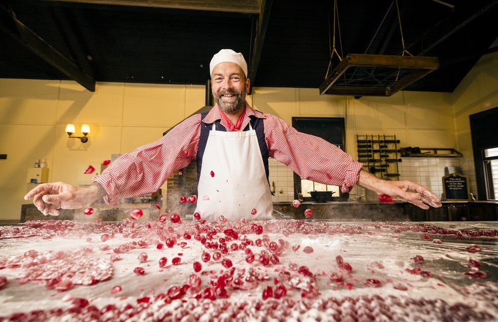 흰 앞치마와 모자를 두른 수염 난 남성이 탁자 뒤에 서서 가루로 덮인 표면에서 빨간 사탕을 다루며 활짝 웃고 있습니다.