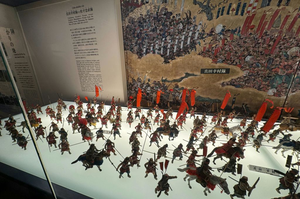 역사적인 전투 예술 작품과 일본어로 된 텍스트 앞에 붉은 깃발이 있는 전투 대형의 미니어처 병사 전시.