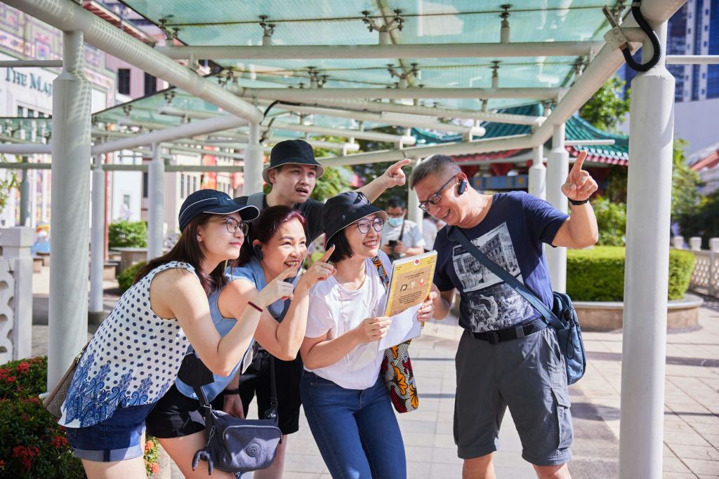 화창한 날, 웃으며 가리키는 다섯 명이 그늘진 야외 구조물 아래에서 종이 지도를 살펴봅니다.