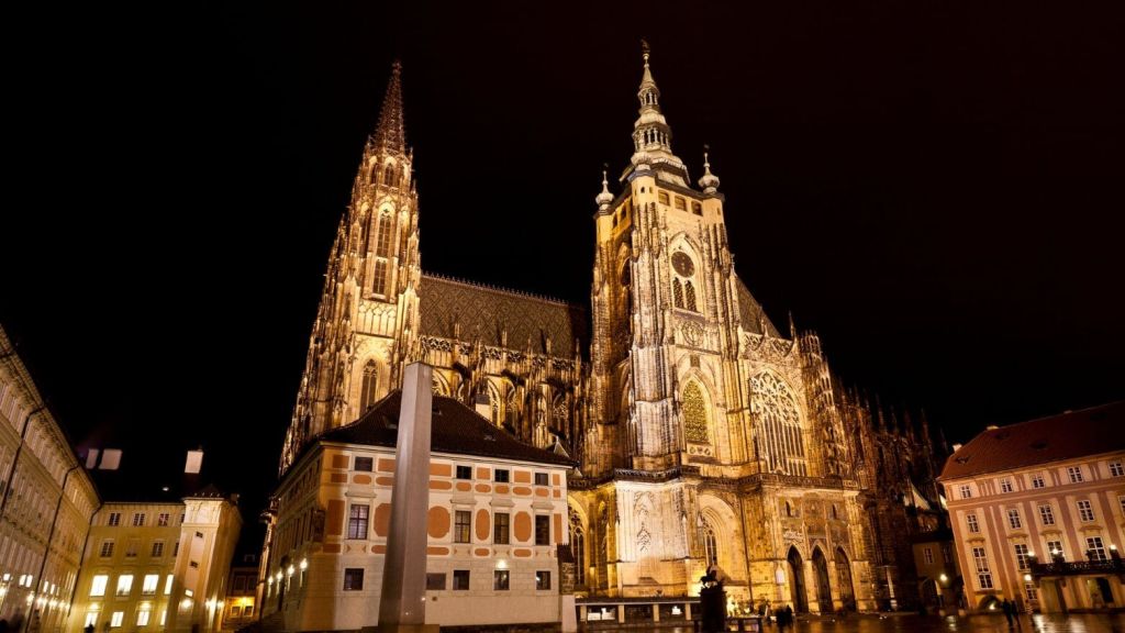 밤에는 정교한 디테일과 높은 첨탑이 있는 크고 빛나는 고딕 양식의 대성당이 전경의 작은 건물과 나란히 있습니다.