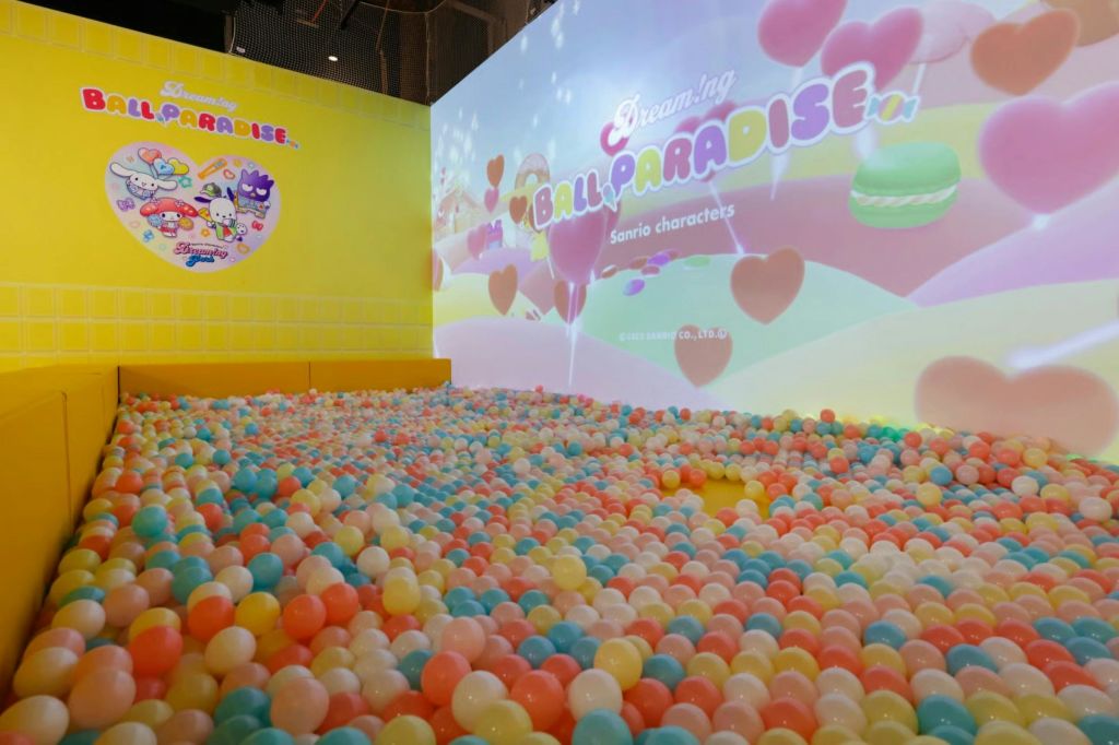 산리오 캐릭터 이미지와 텍스트가 표시된 벽 앞에 파스텔 색상의 볼이 있는 다채로운 볼 피트가 있습니다.