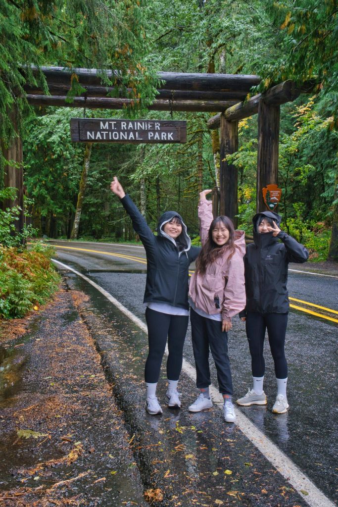 산 아래 야외 장비를 착용한 세 사람이 서 있습니다. 숲이 우거진 도로에 있는 레이니어 국립공원 표지판에서 미소를 짓고 포즈를 취하고 있습니다.